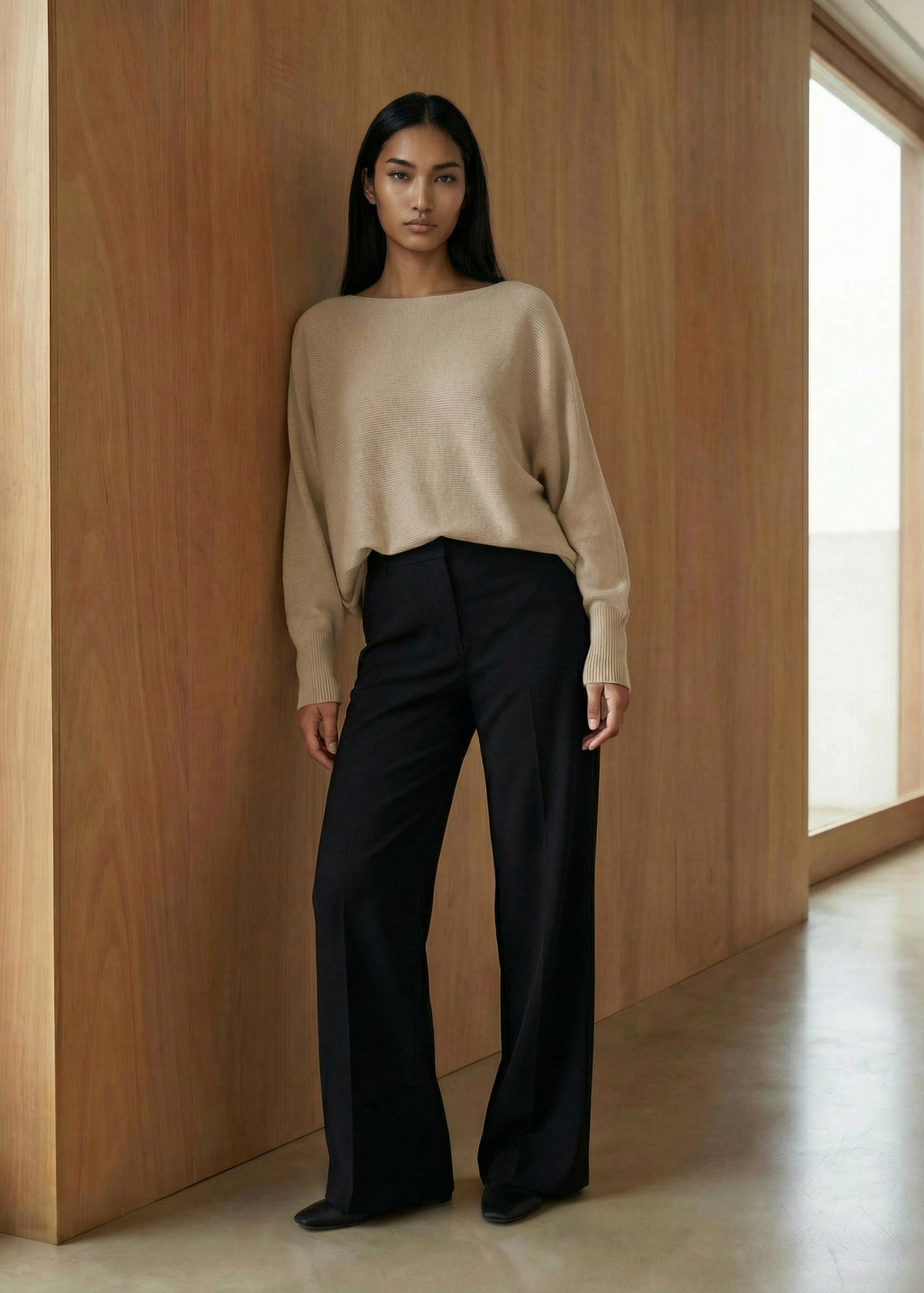 Woman wearing a beige sweater and black pants in a minimalistic room with wooden walls and a large window.
