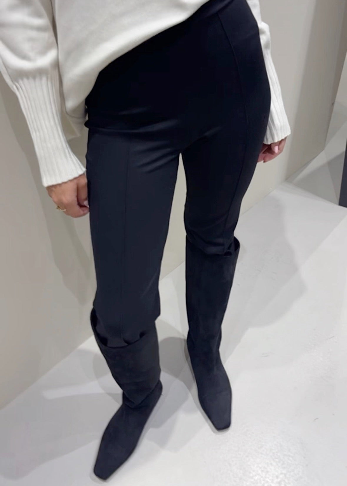 Person wearing navy blue knee-high boots and a white sweater against a plain background