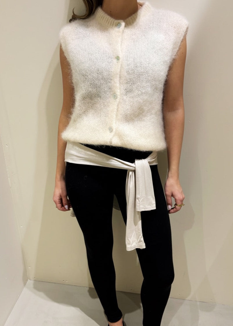 Person wearing a white knitted vest over a black outfit against a plain background