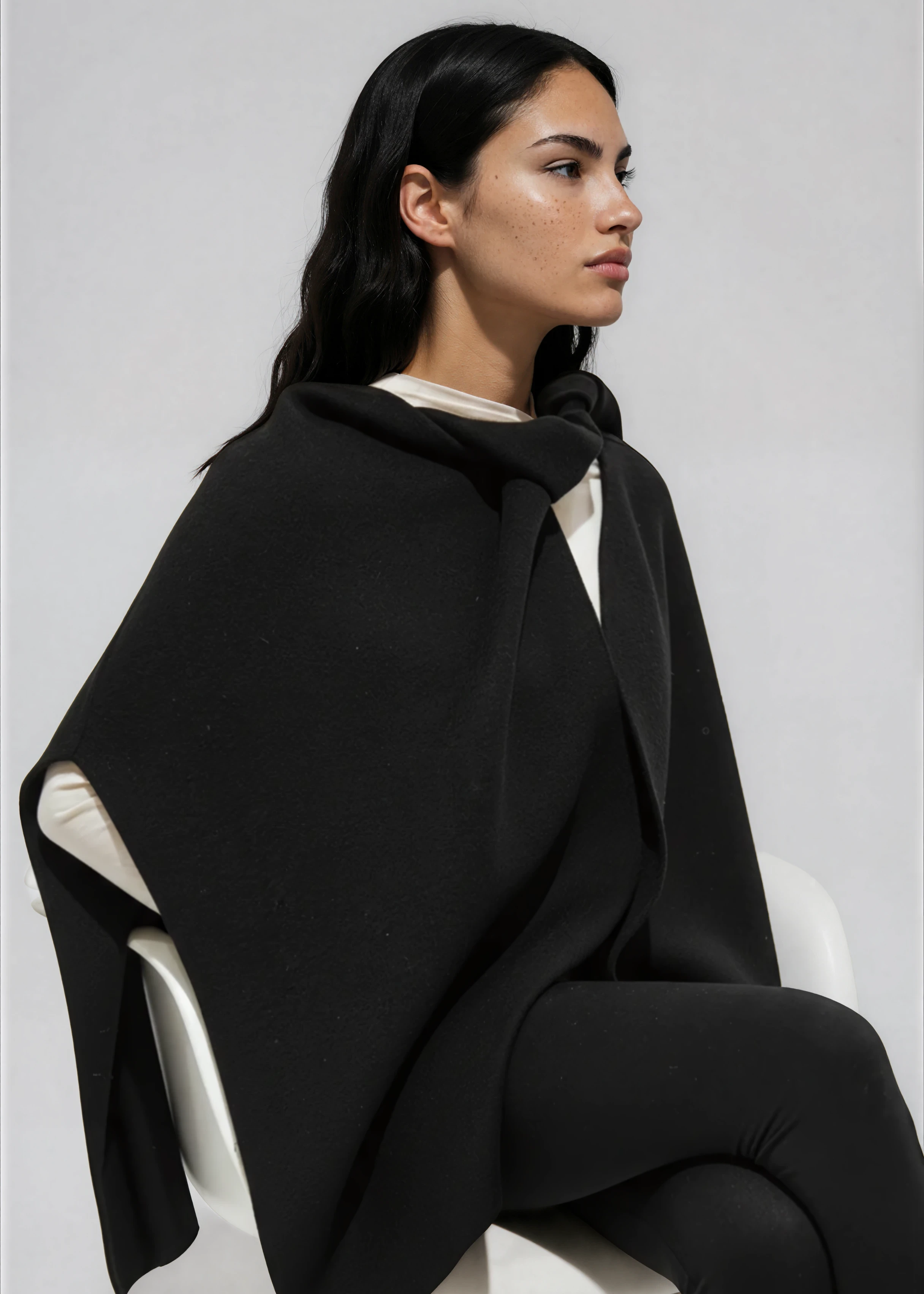 Woman wearing a black poncho sitting on a white chair against a light gray background