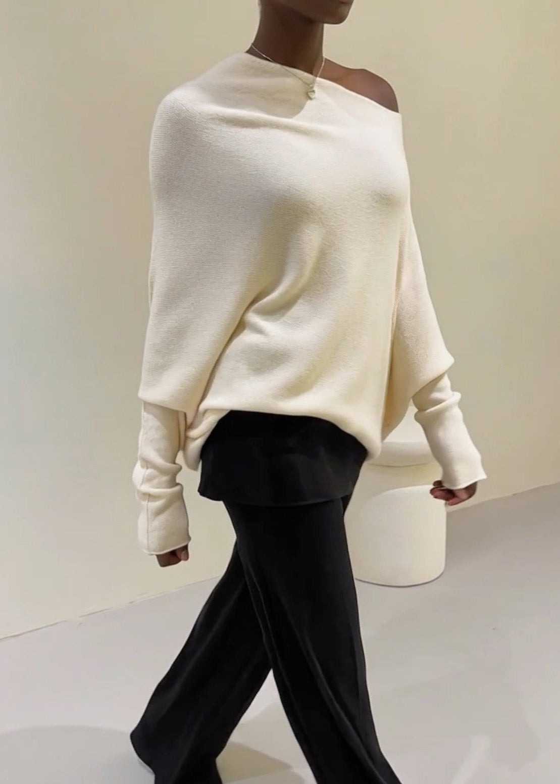 Person wearing a white off-shoulder sweater and black pants on a plain background