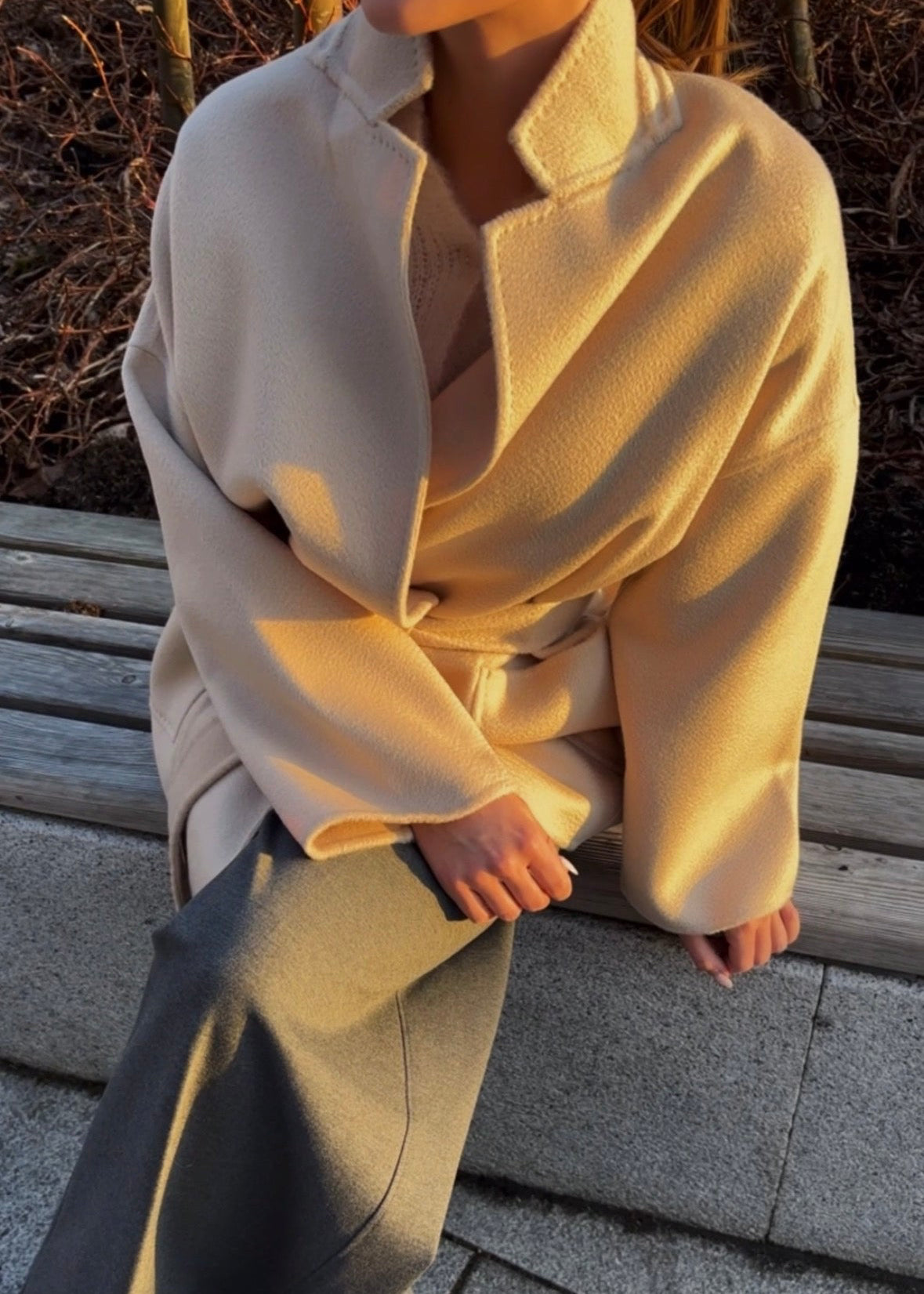 Person wearing a long beige coat sitting on a bench.