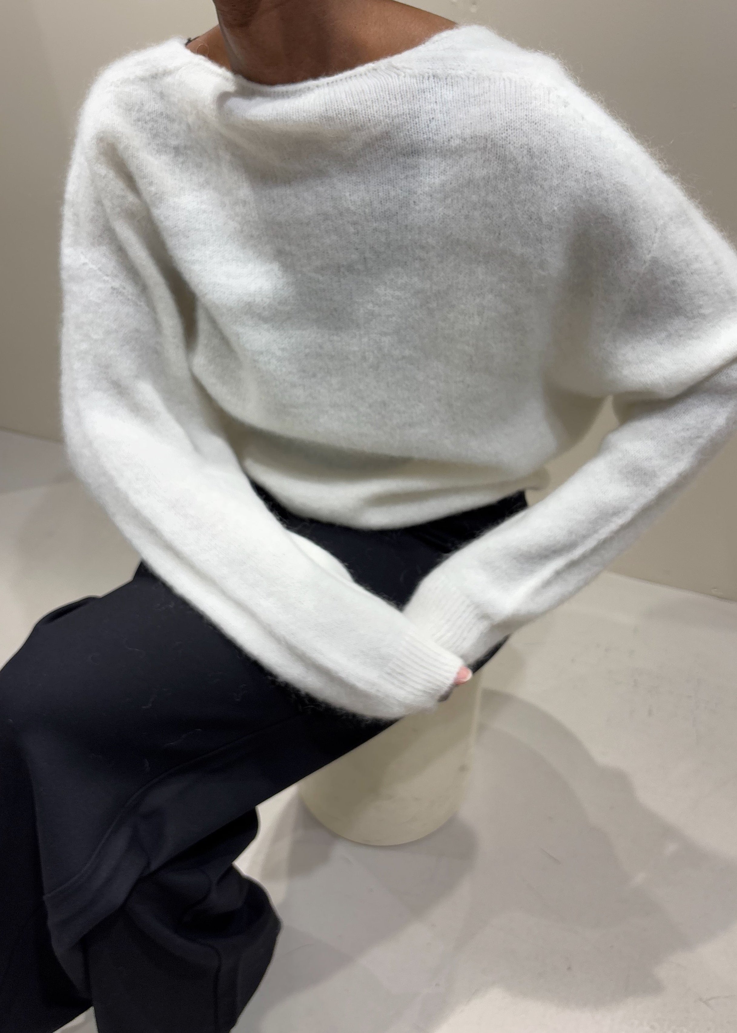 Person wearing a white sweater and dark pants sitting on a white stool against a plain background