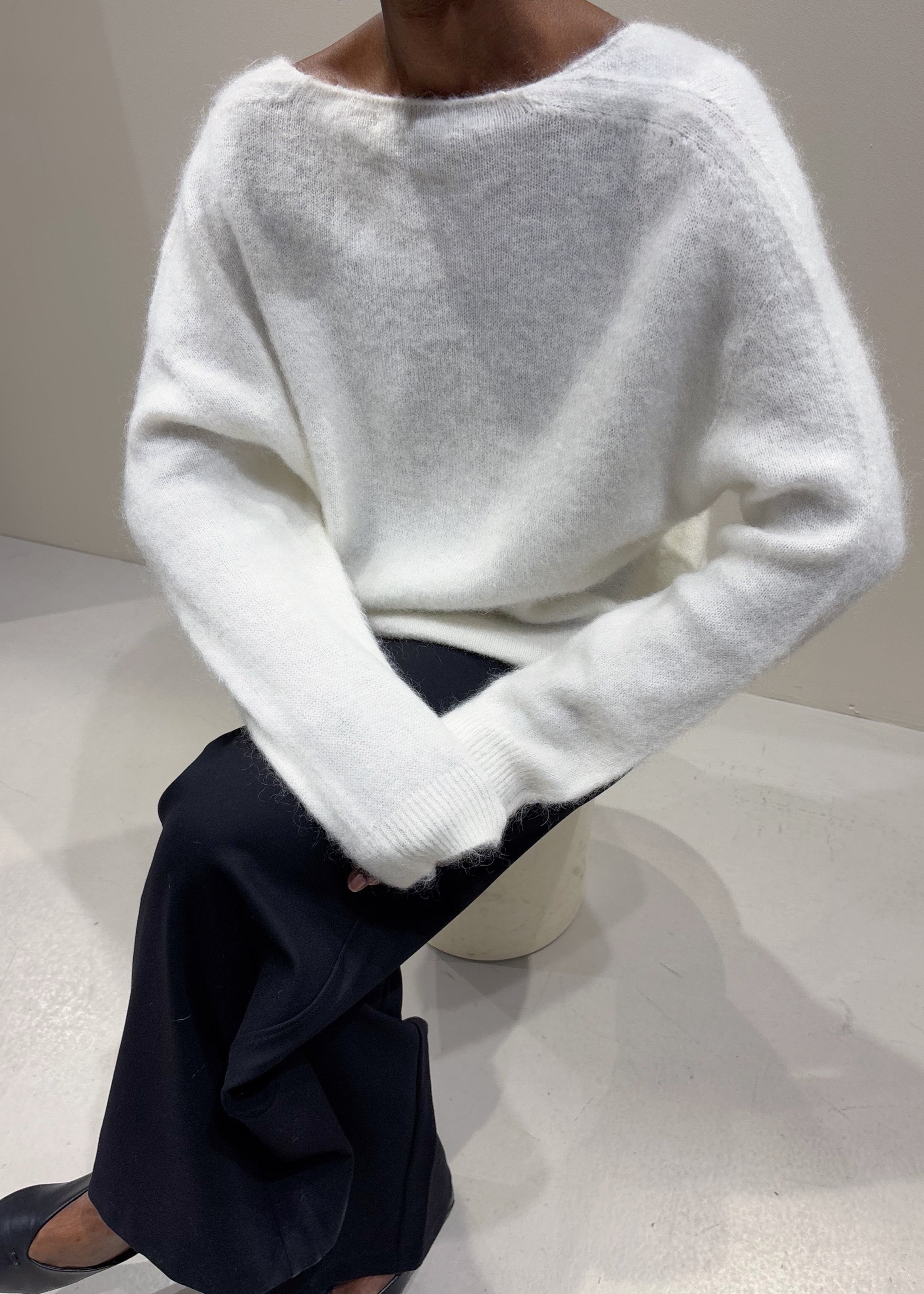 Person wearing a white sweater and dark pants sitting on a white surface.
