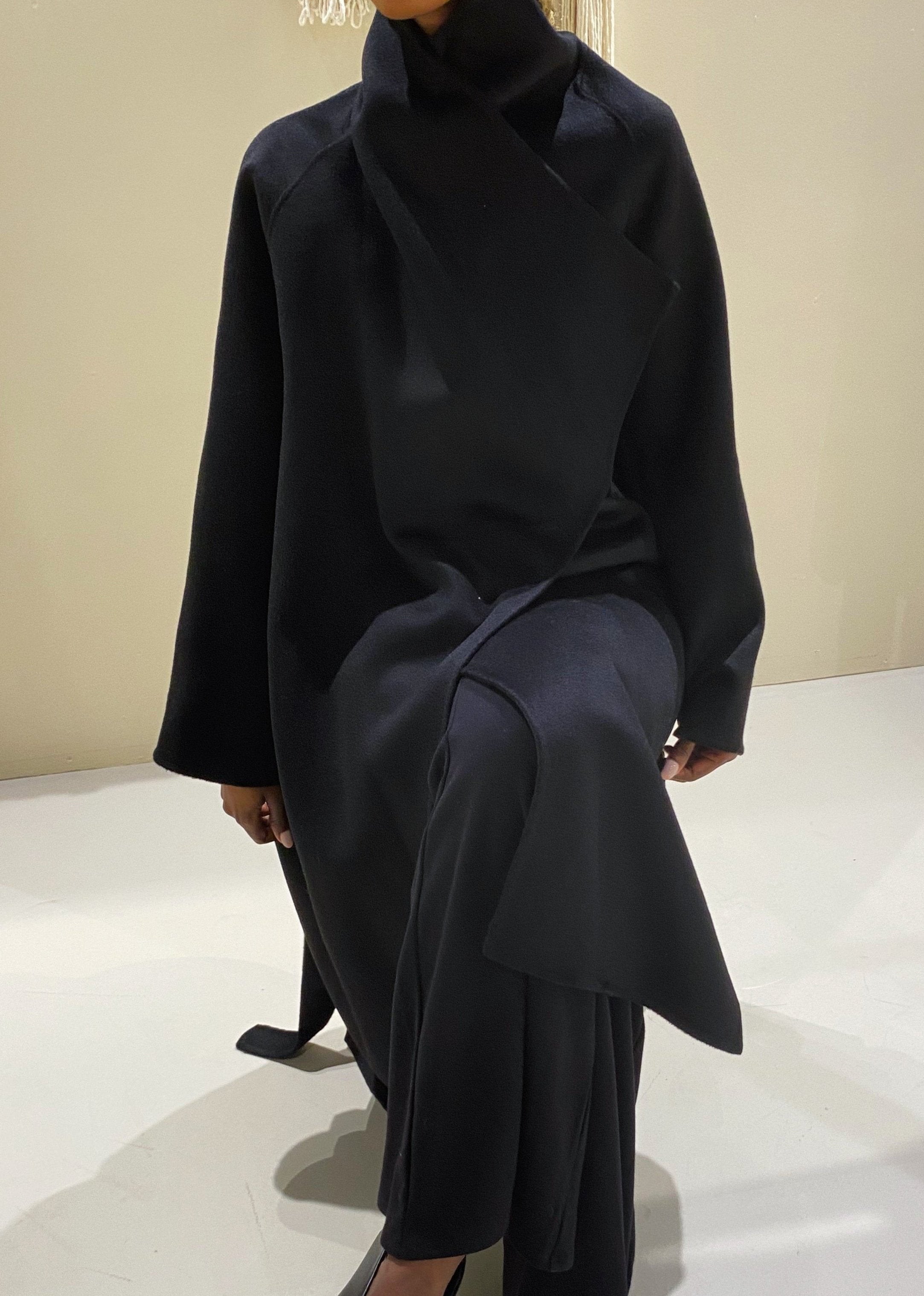 A person wearing a long, belted, black wrap coat with exaggerated lapels, sitting in a relaxed pose.