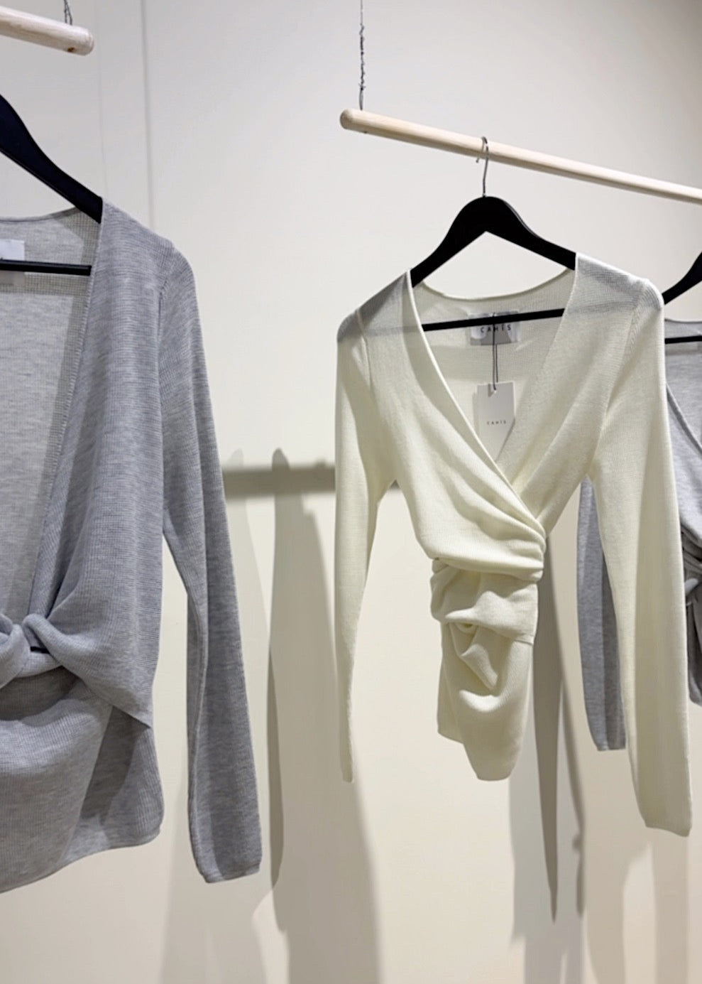 White long-sleeve top with a V-neck and knot detail on a hanger against a white wall.