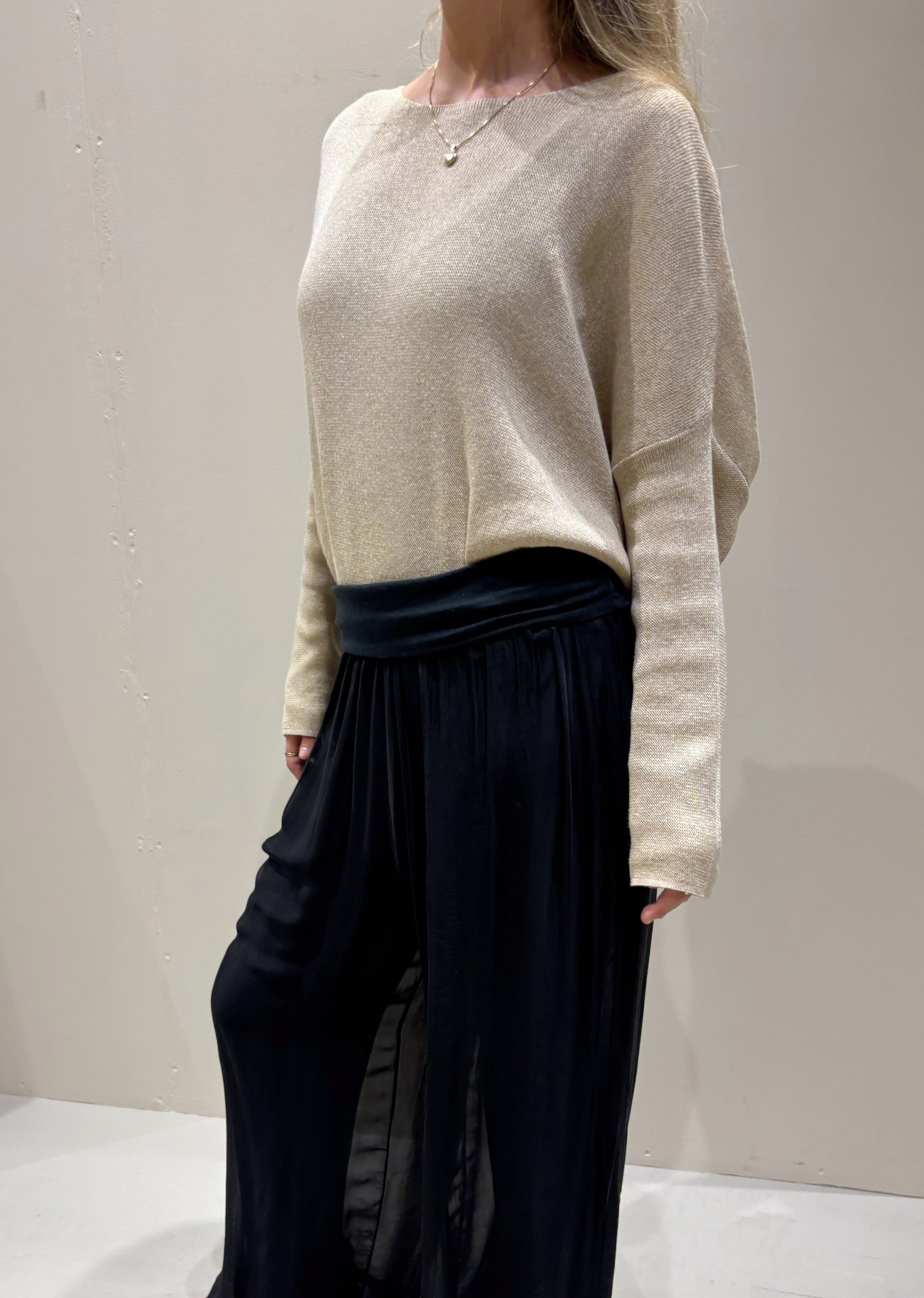 Person wearing a beige sweater and black pleated pants against a plain background