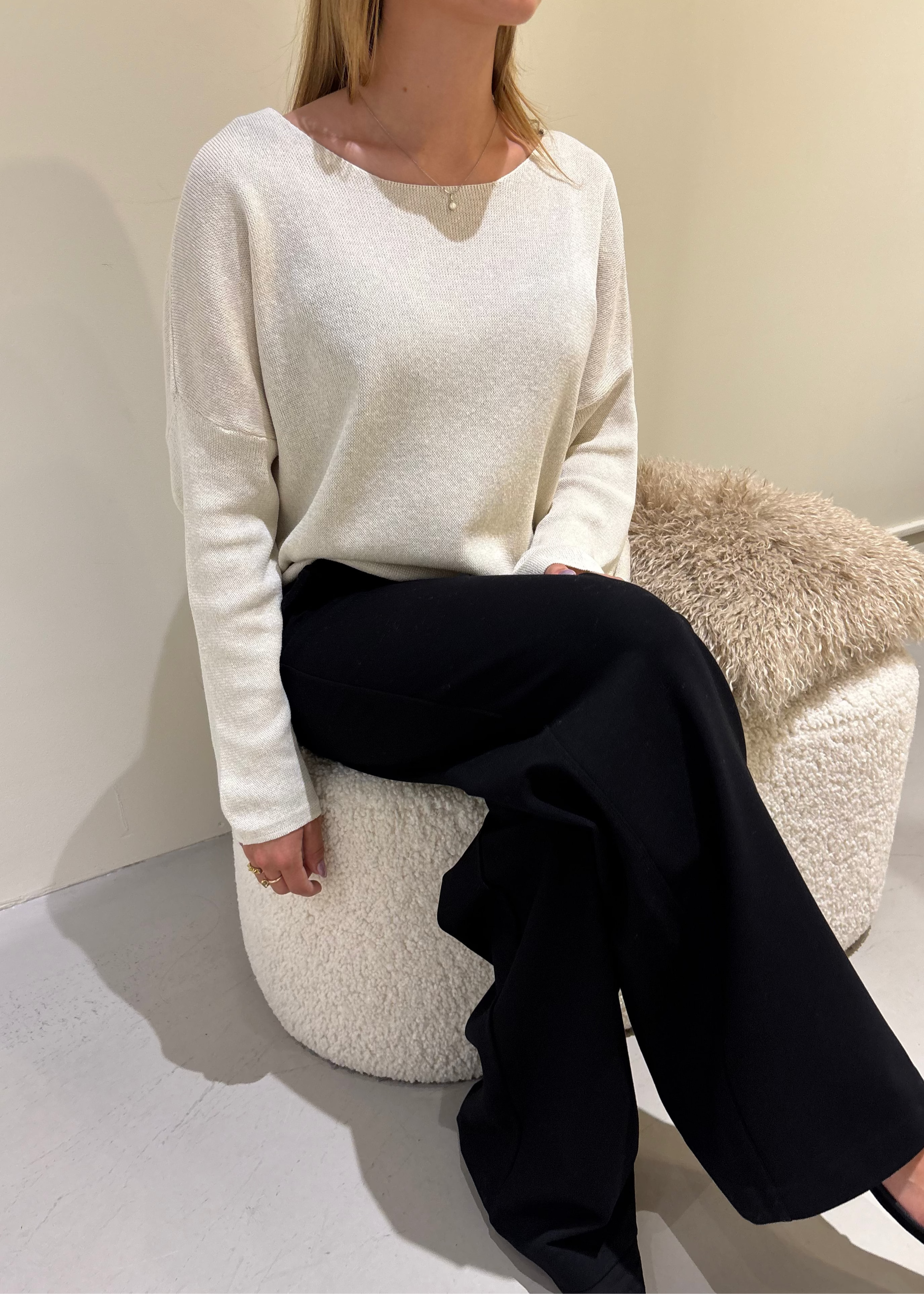 Person wearing a white sweater and black pants sitting on a textured stool against a plain wall.
