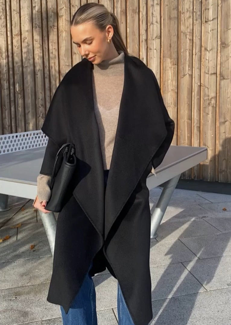 A person wearing a belted, long silhouette black Talori wrap coat with exaggerated lapels and side slits, over a beige turtleneck, paired with blue jeans.