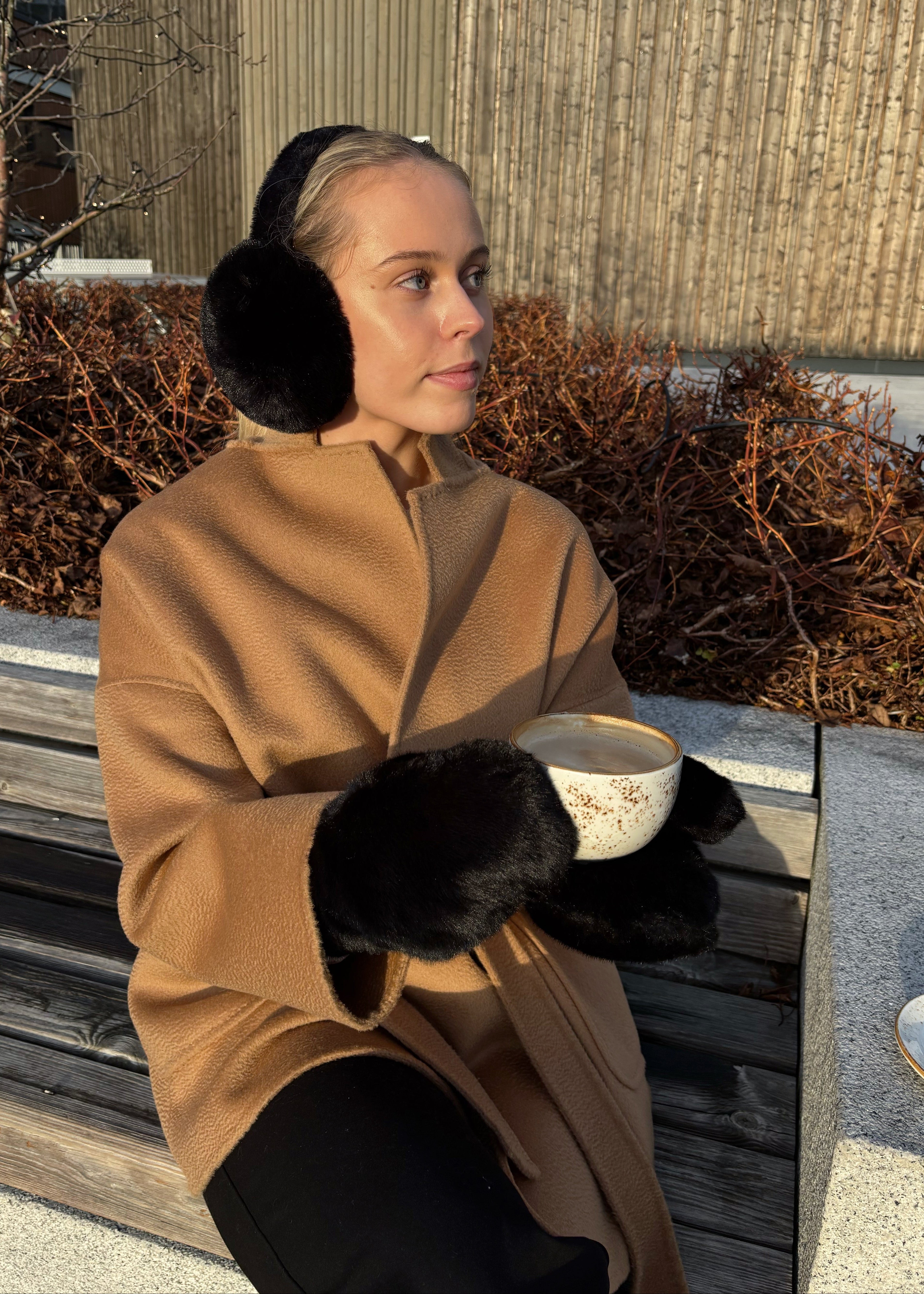 Woman in a brown coat and black earmuffs sitting on a bench with a cup.