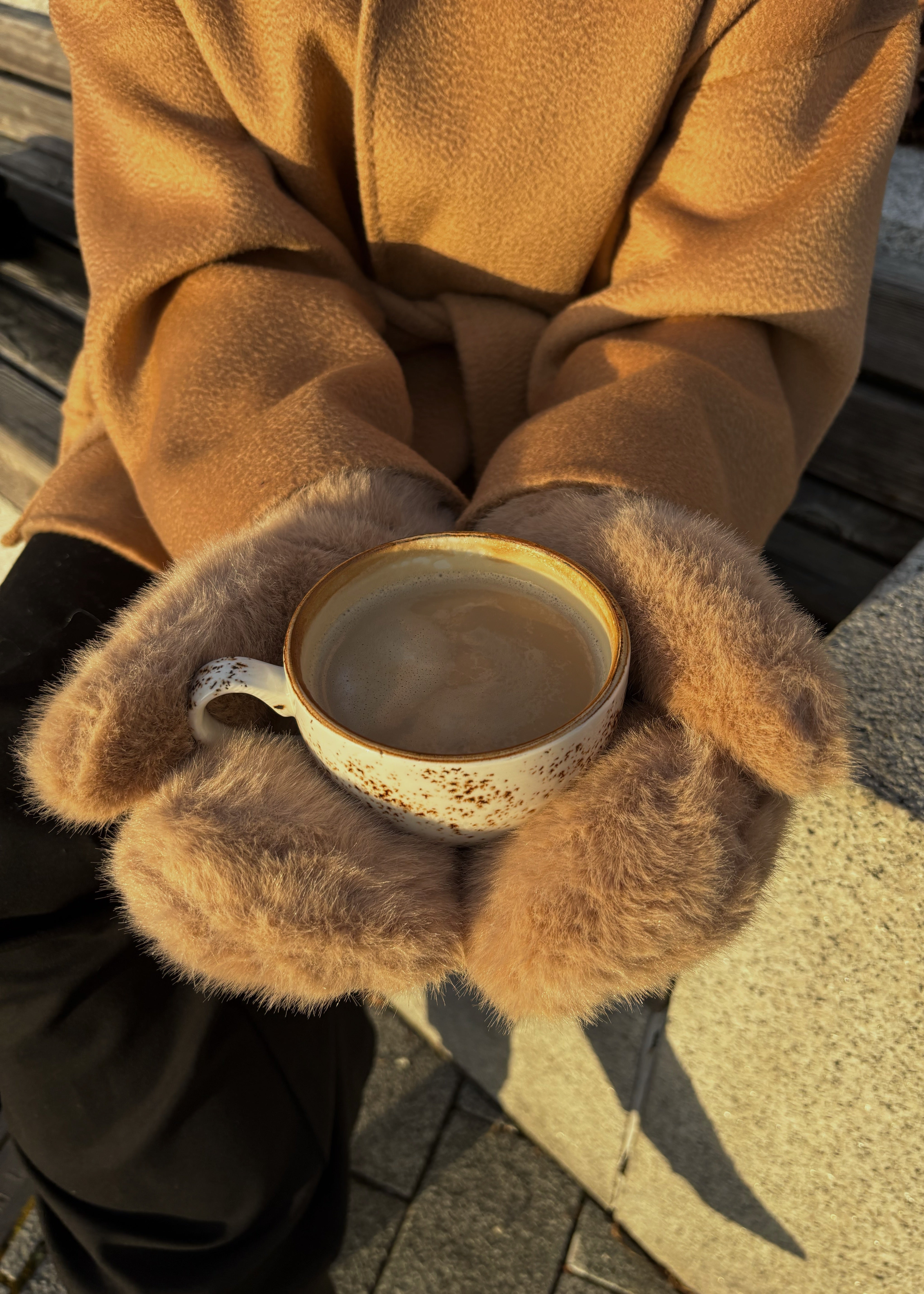 Person wearing a brown coat and fur-lined gloves holding a cup outdoors.