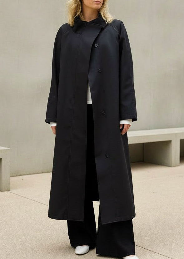 Woman wearing a long black coat in an indoor setting with concrete walls and benches.