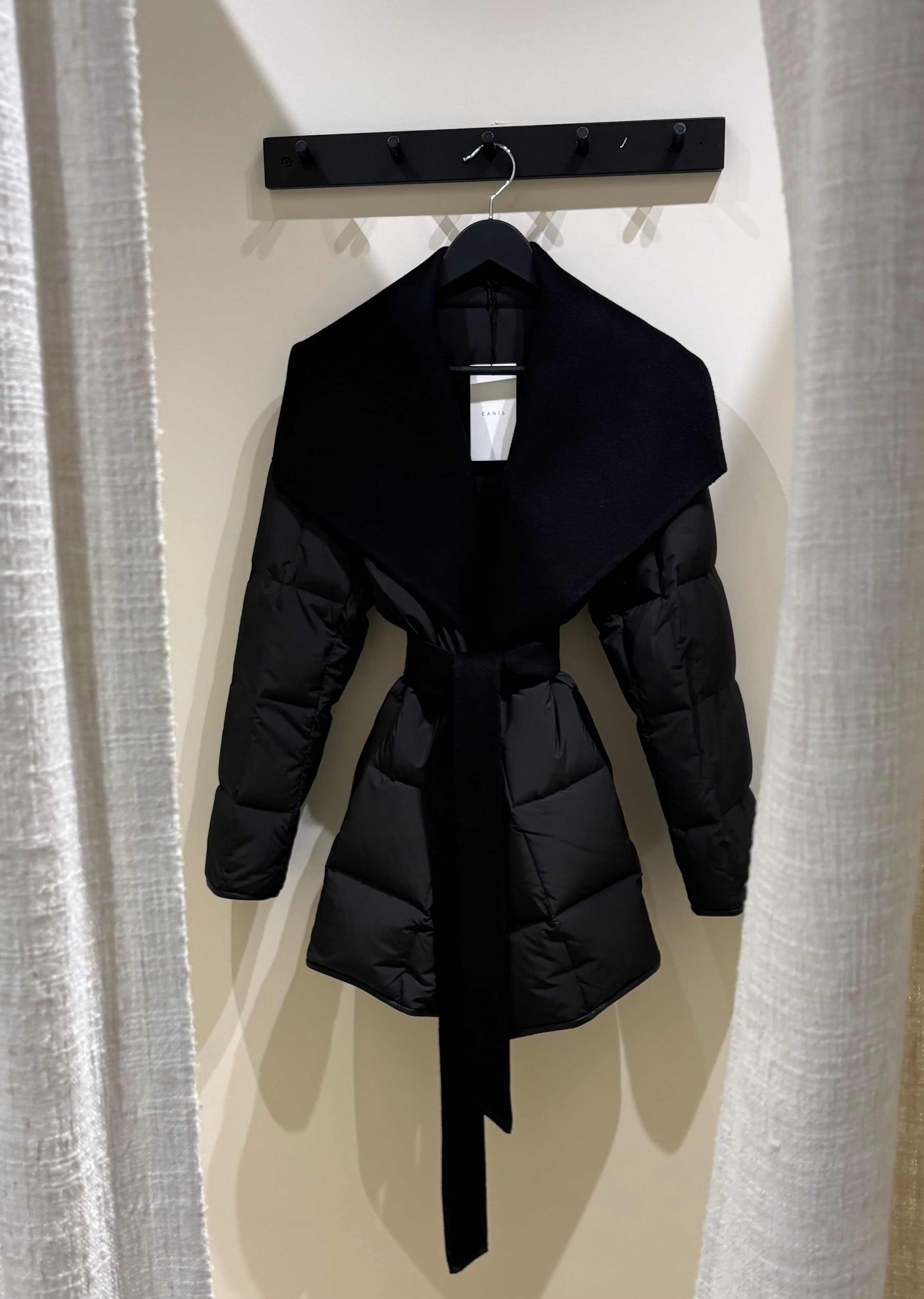 Black puffer coat with a belt hanging on a rack between two curtains.