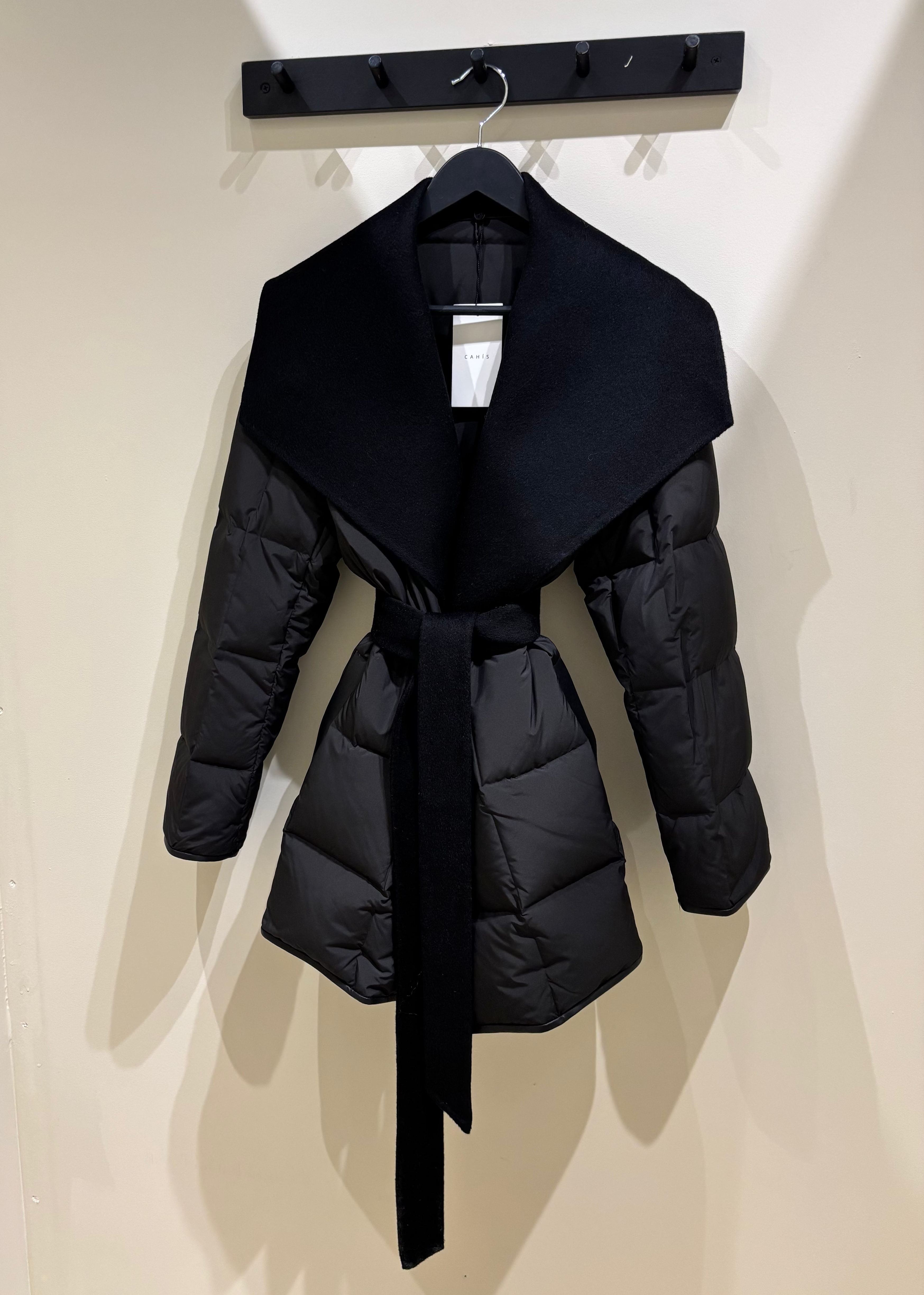 Black puffer jacket with a belt hanging on a rack against a white wall.