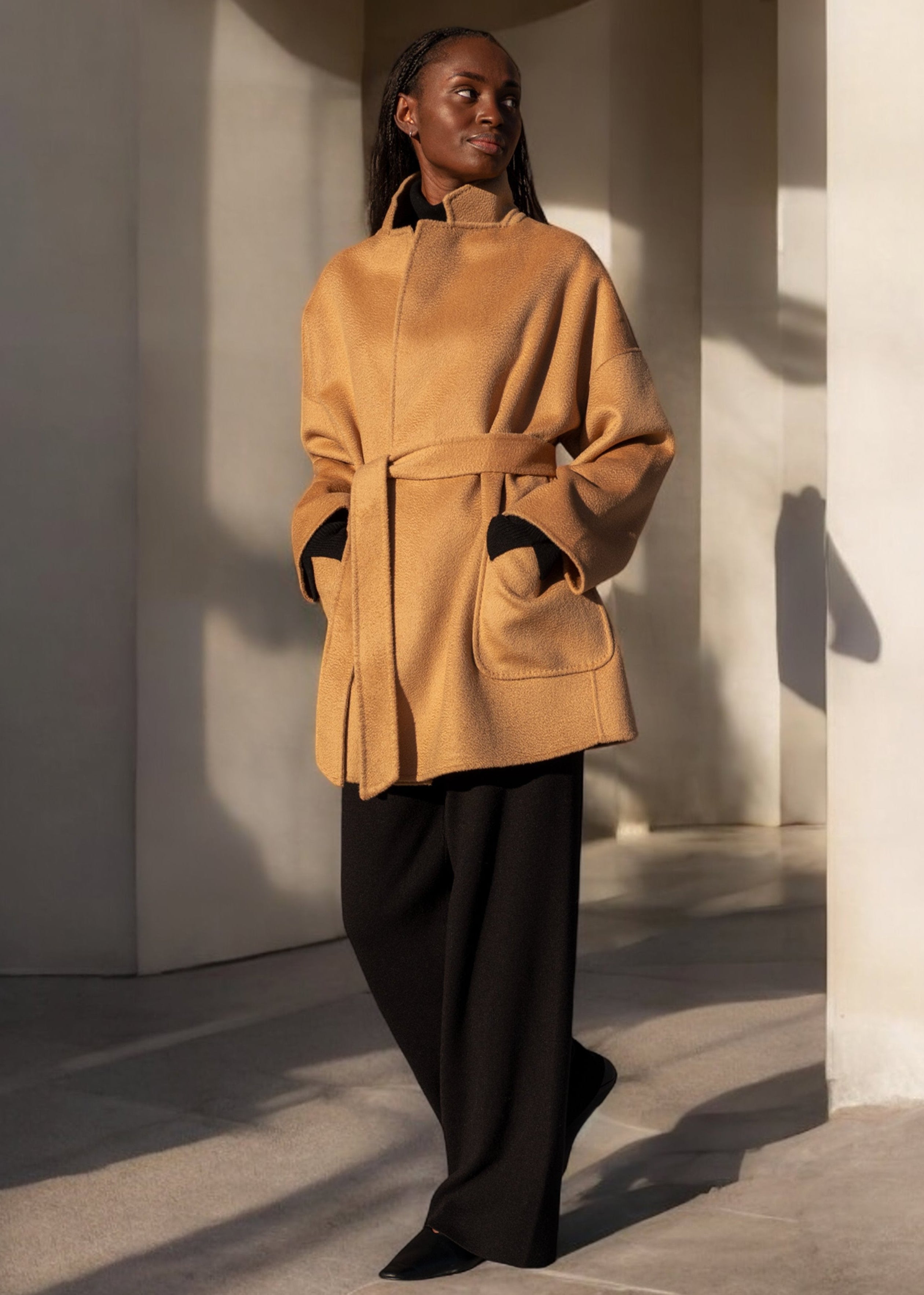 A person standing in a relaxed pose wearing a beige double face wool jacket with a stand collar and deep patch pockets, fastened with a matching belt.