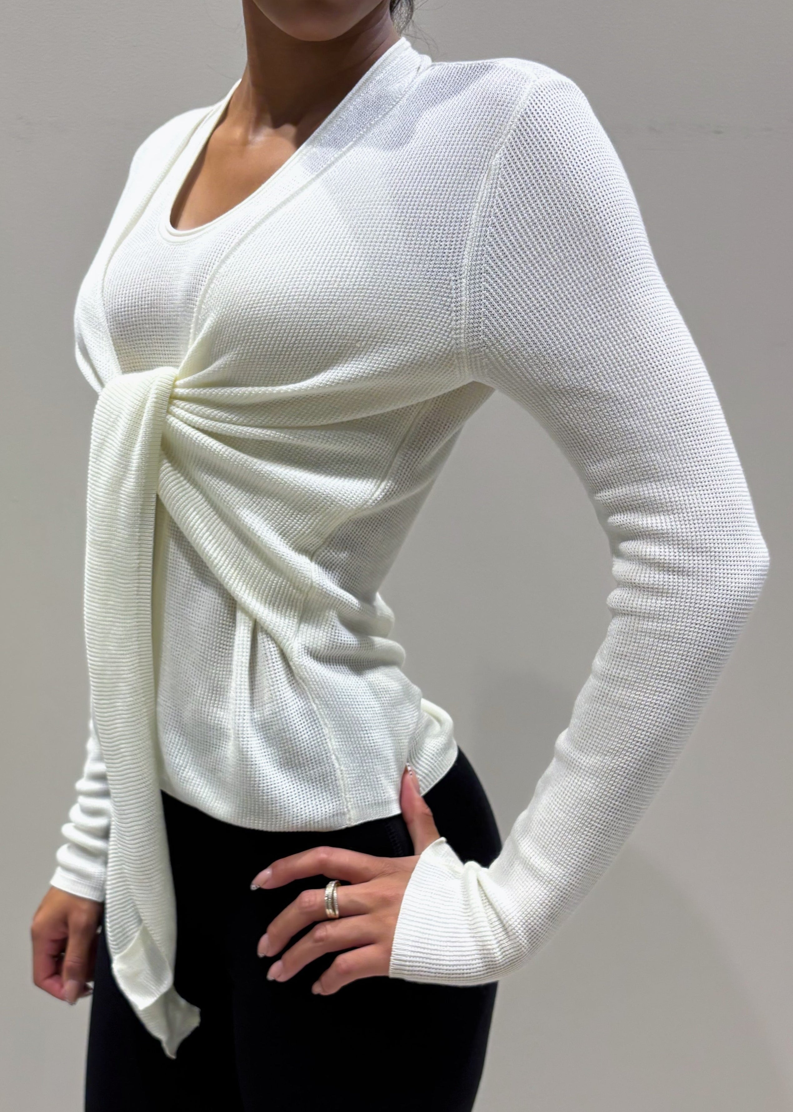 Person wearing a white long-sleeve top with a knot detail and black pants against a plain background