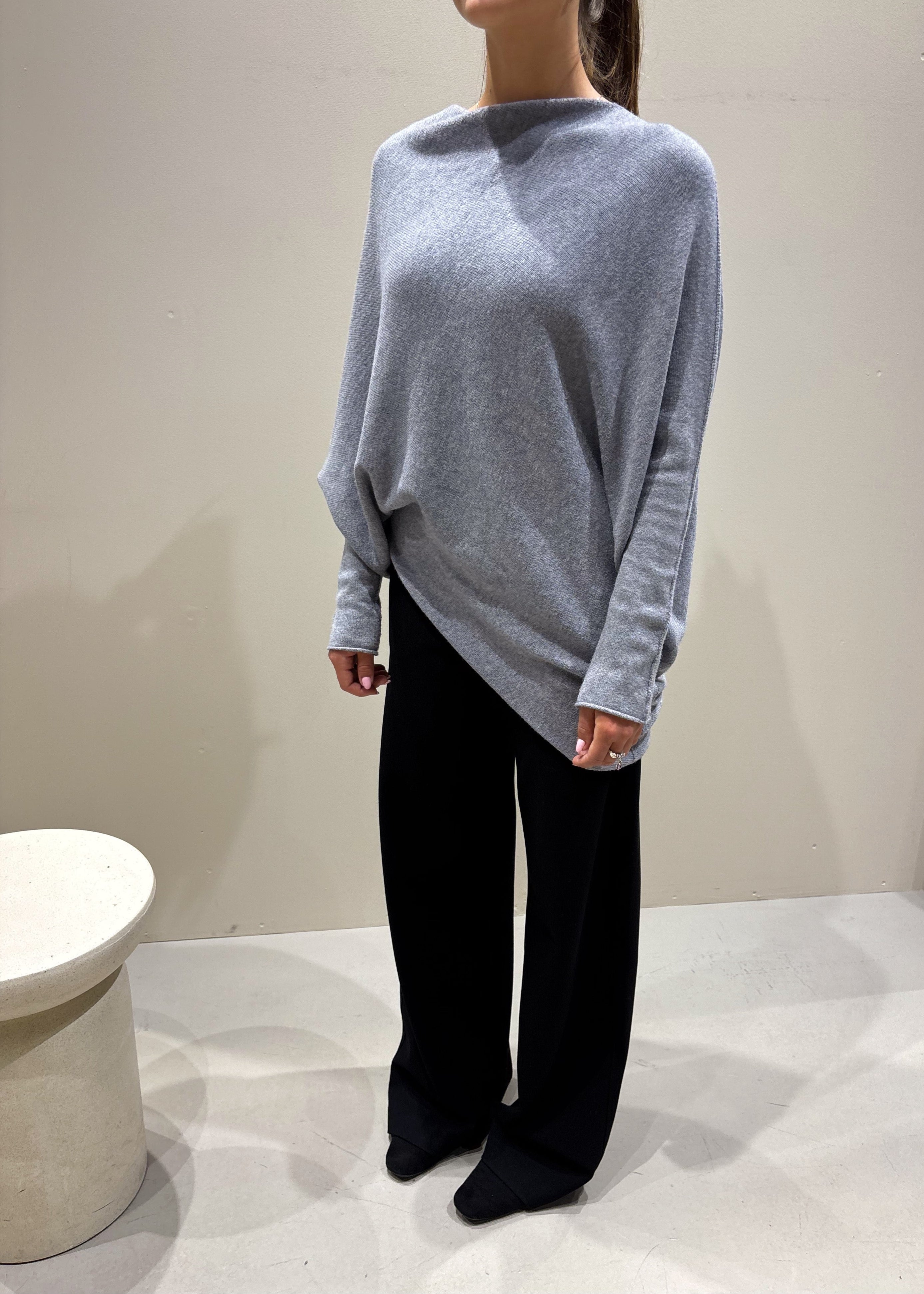 Person wearing a light gray sweater and black pants standing against a plain background