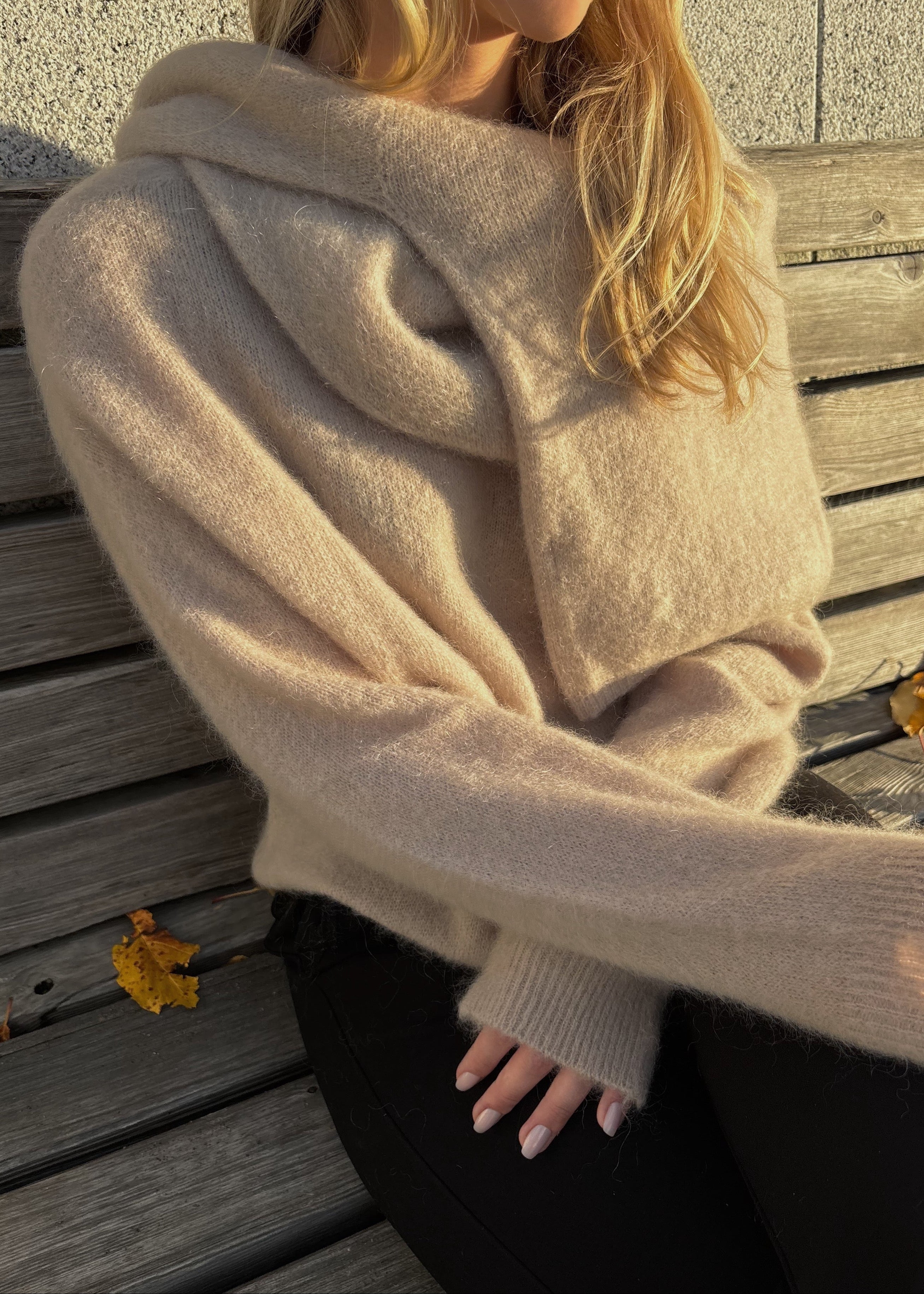 Person wearing a beige sweater sitting on a wooden bench with autumn leaves.