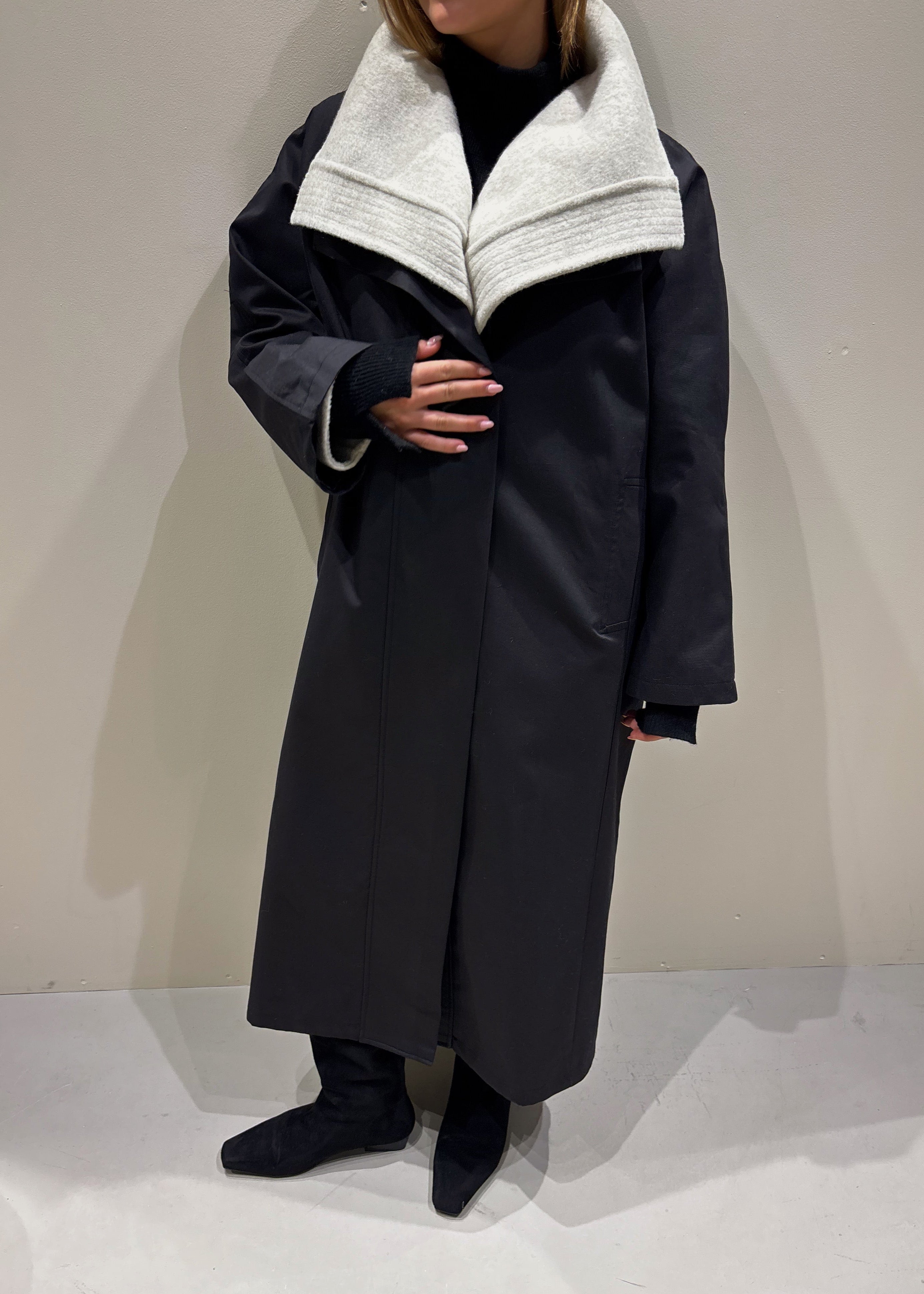 Person wearing a long dark coat with a light gray collar against a plain background