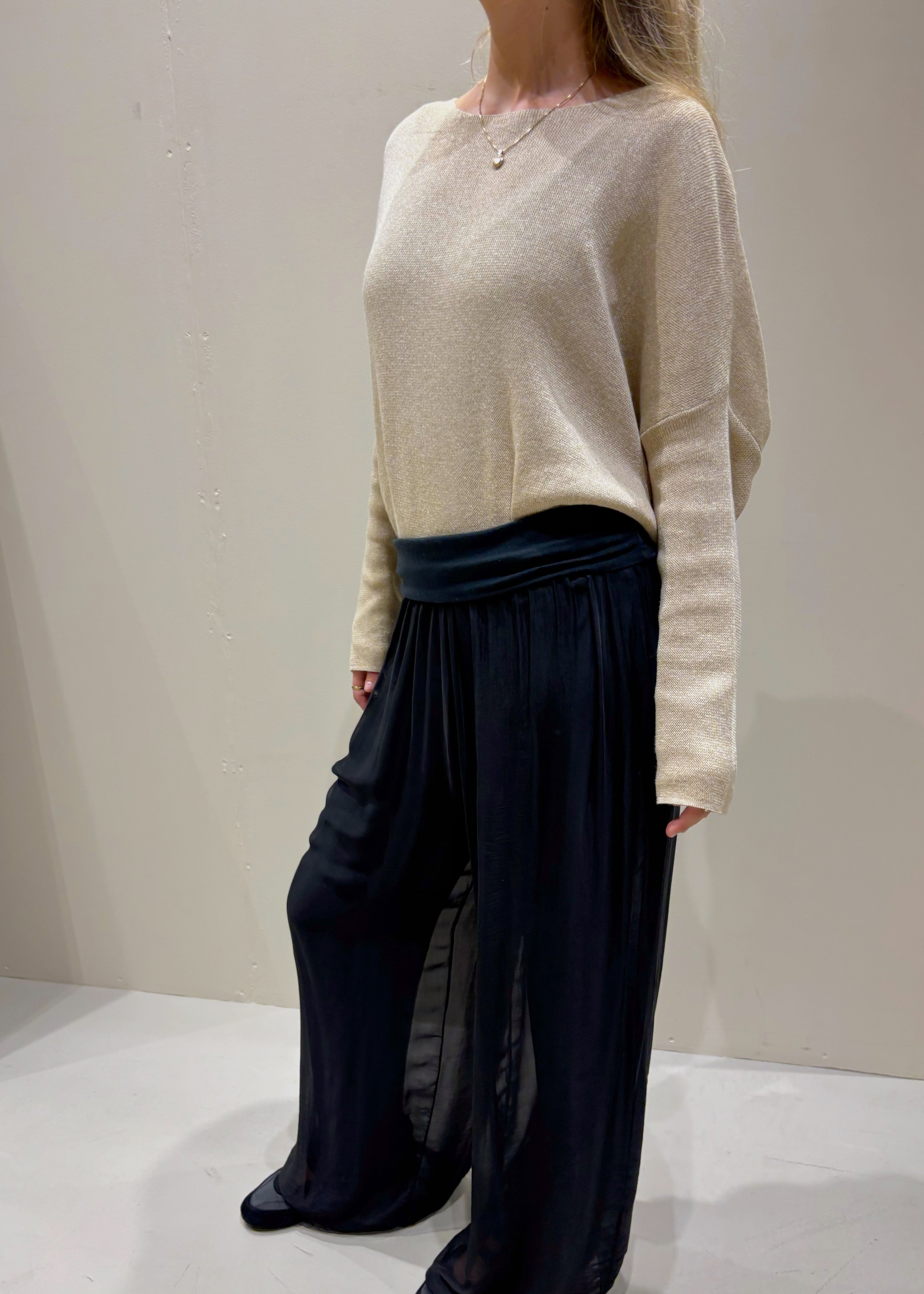 Person wearing a beige sweater and dark pleated pants against a plain background