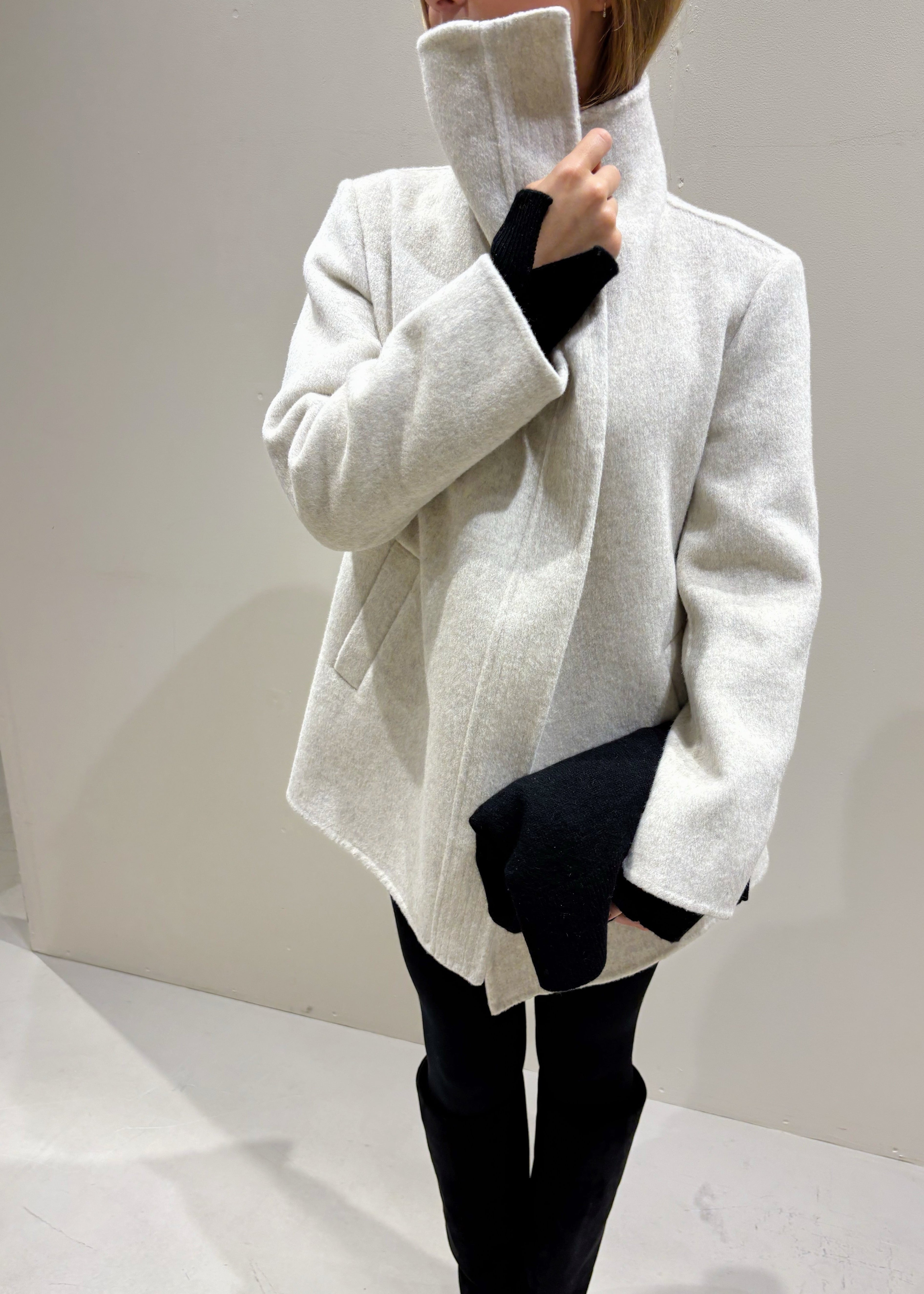 Person wearing a light gray coat over a black outfit on a plain background