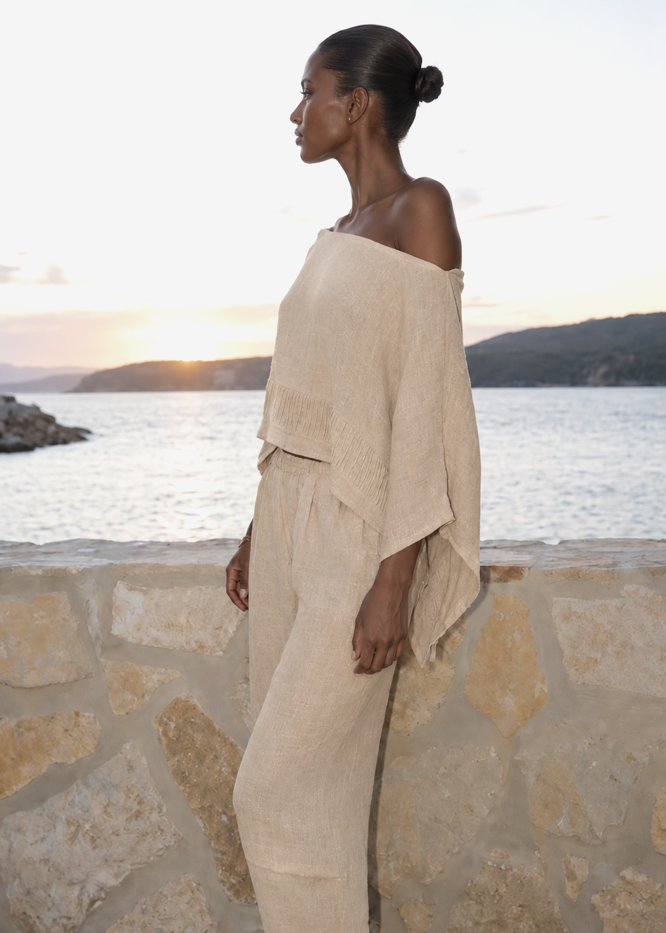 Woman in beige outfit standing by a stone wall with a scenic background
