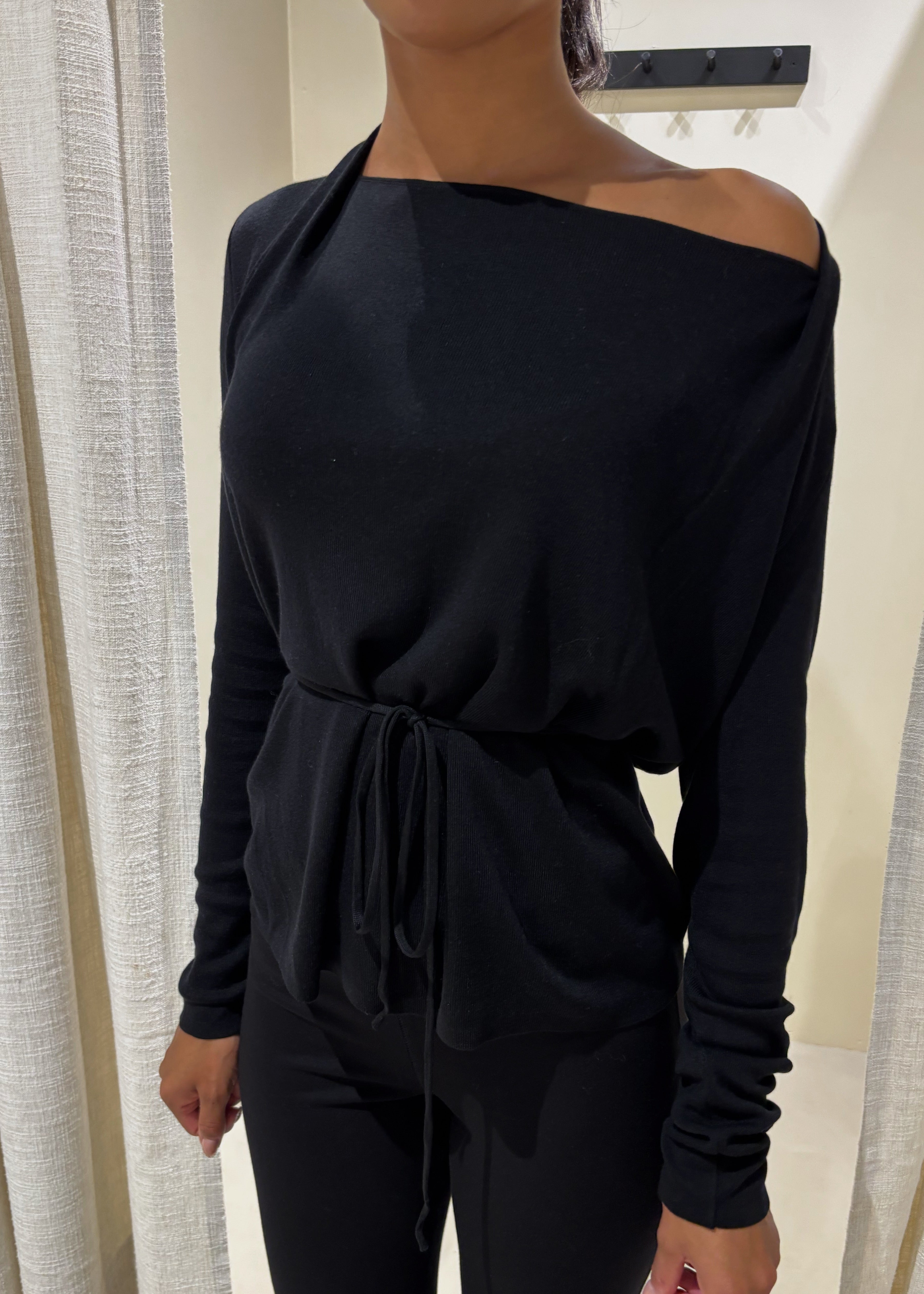 Person wearing a black off-shoulder top and pants in front of white curtains.