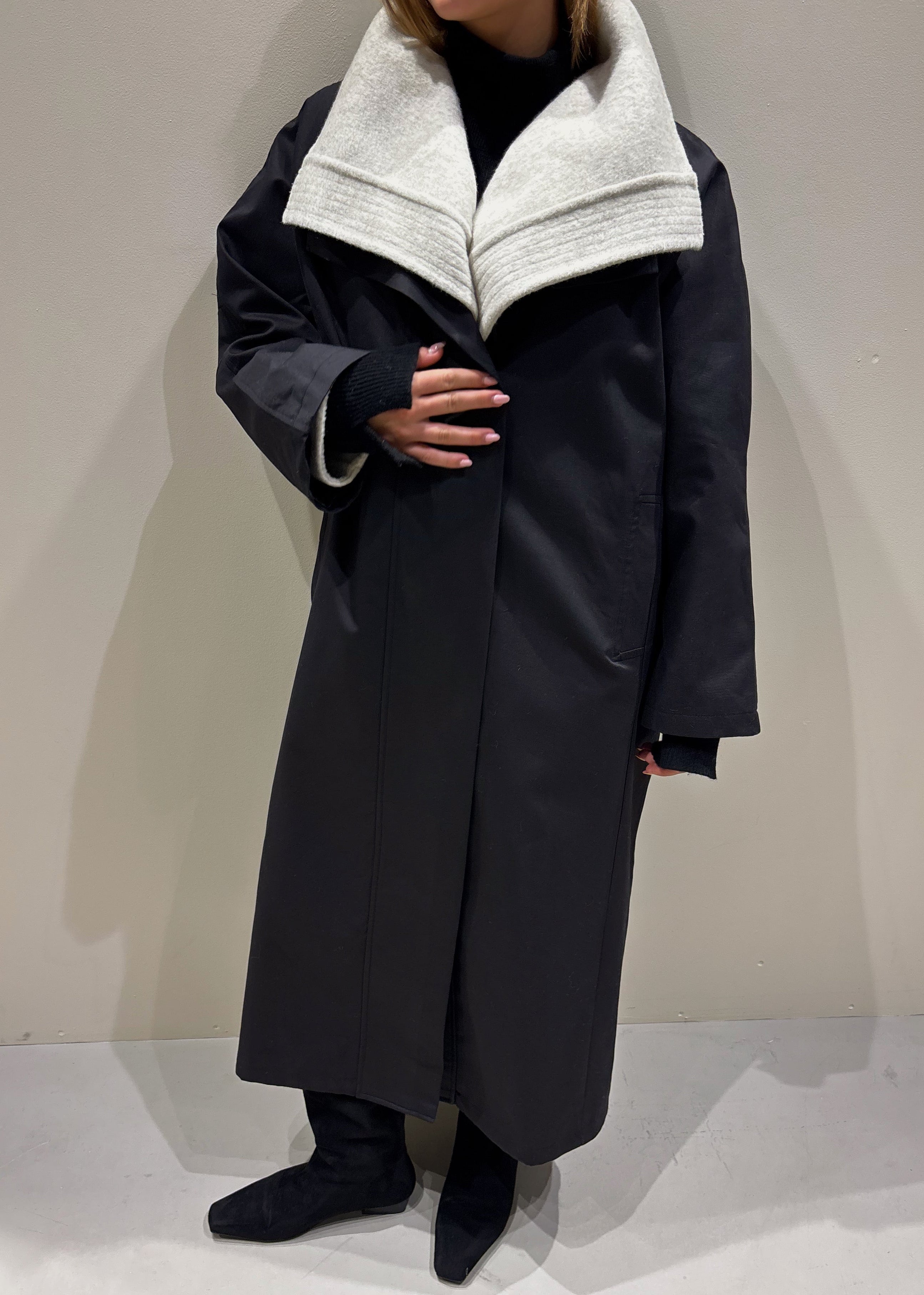 Person wearing a long dark coat with a light gray collar against a plain background