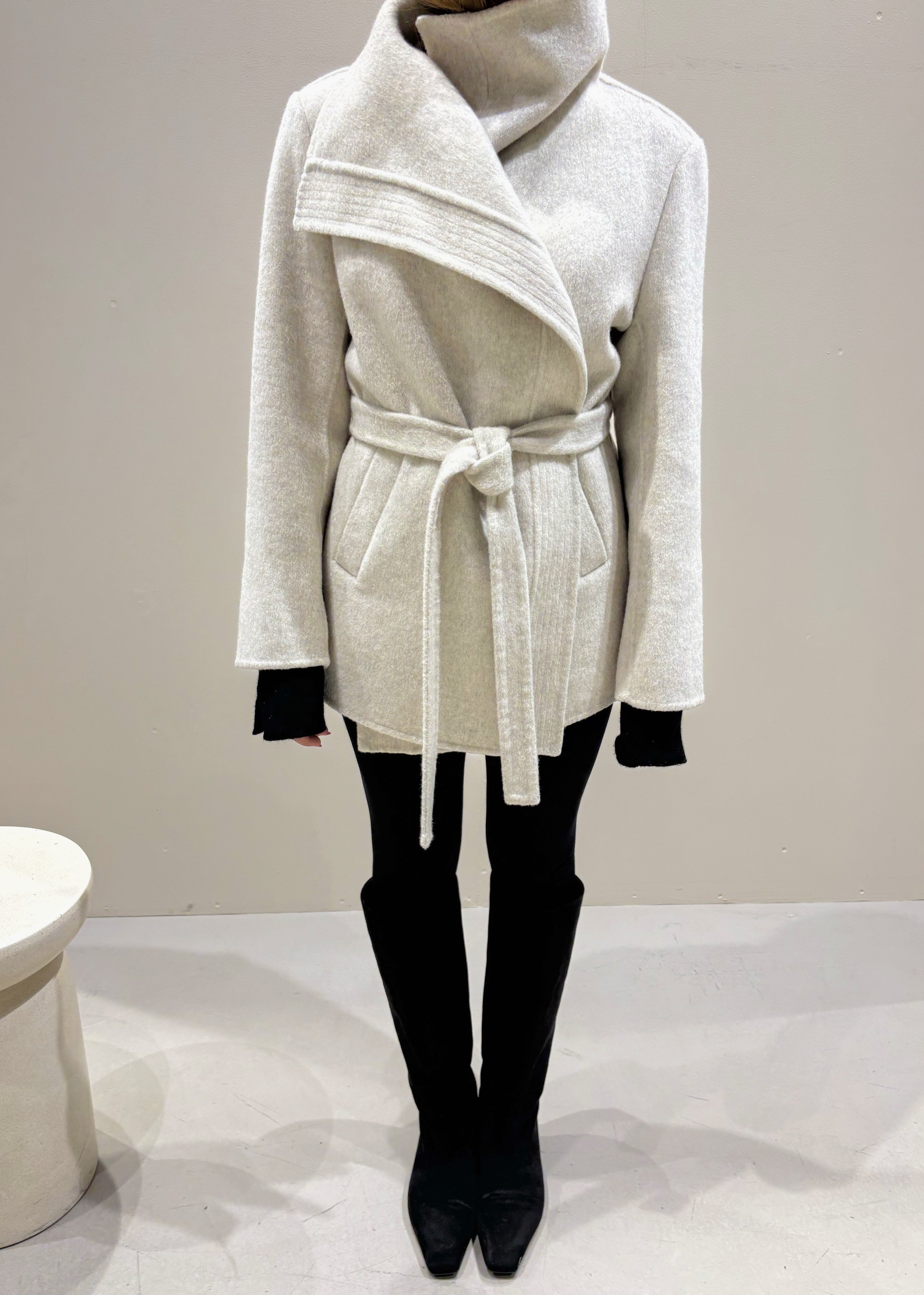 Person wearing a light gray coat with a belt on a plain background