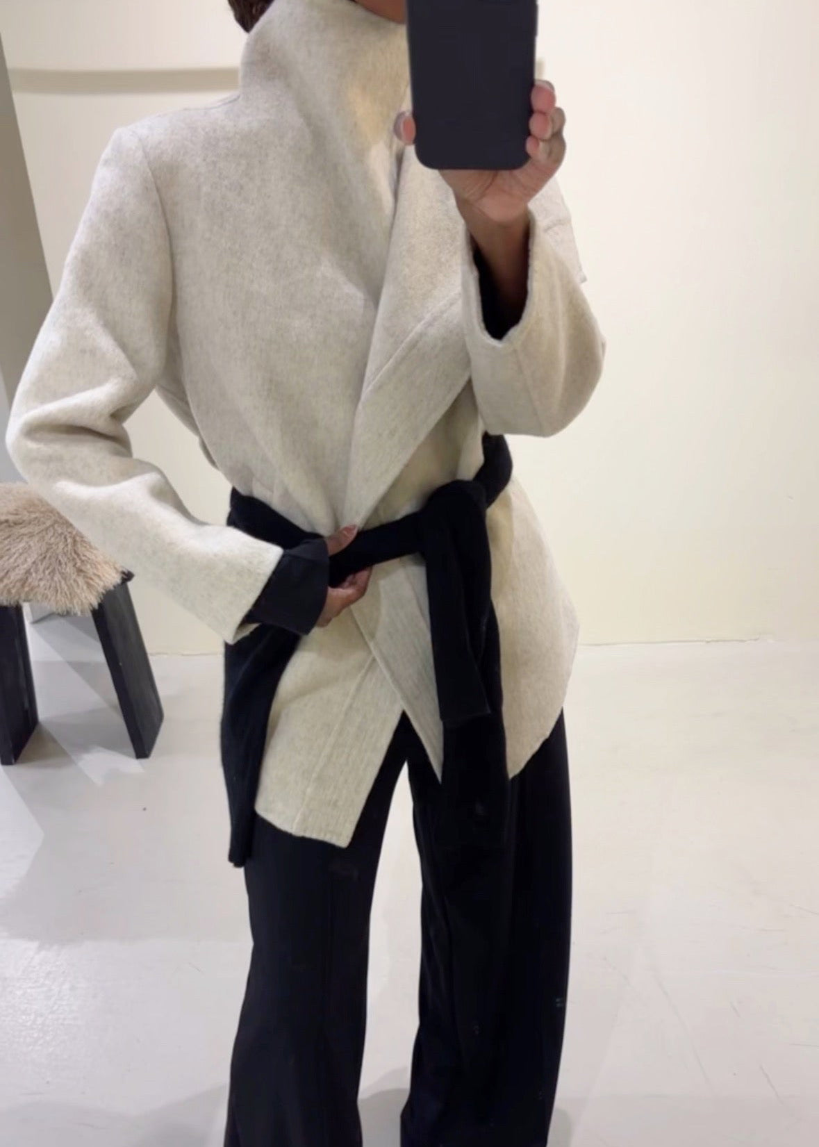 Person wearing a light gray coat with a black belt in a minimal indoor setting.