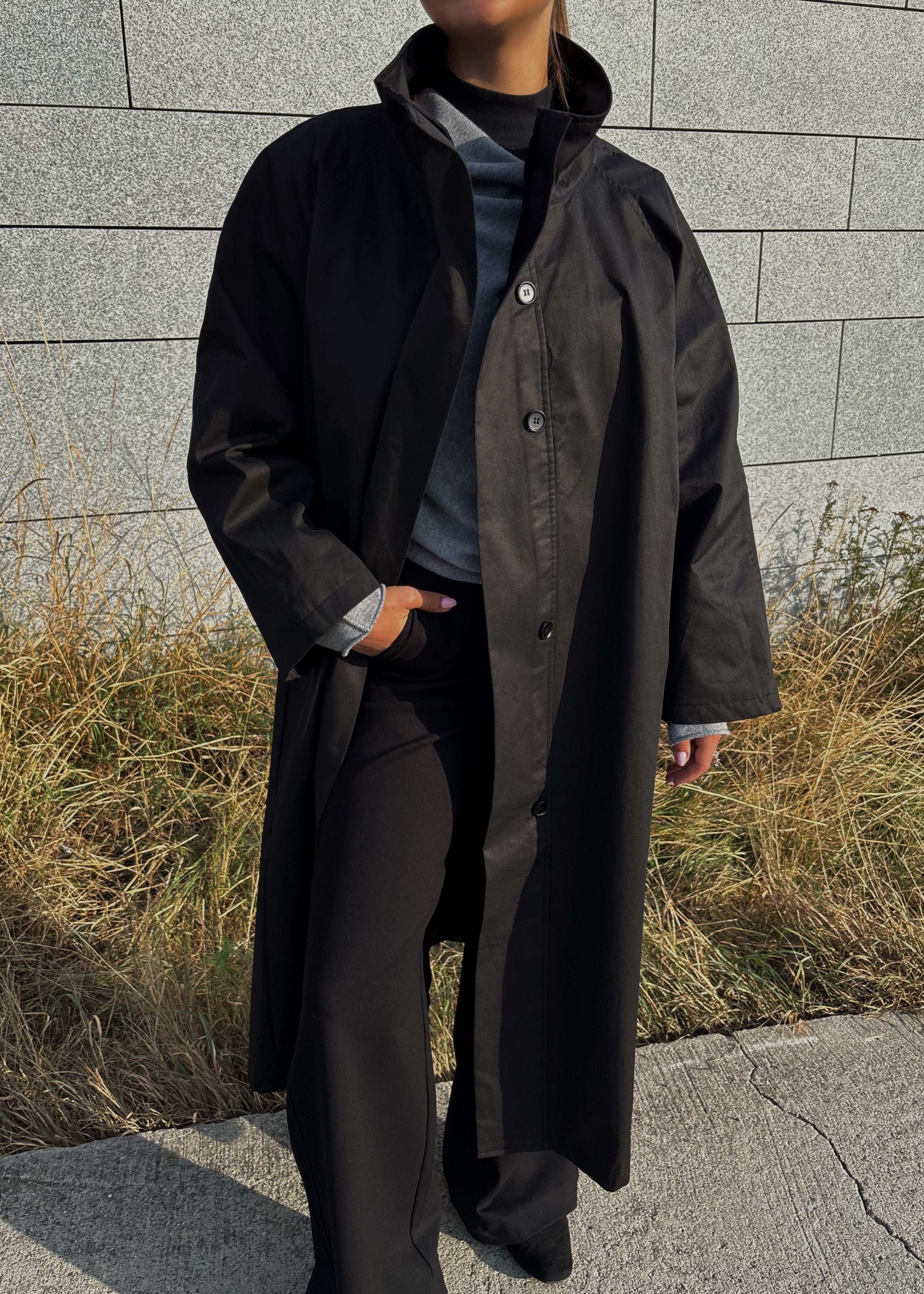 Person wearing a long black coat standing against a gray wall with dry grass.