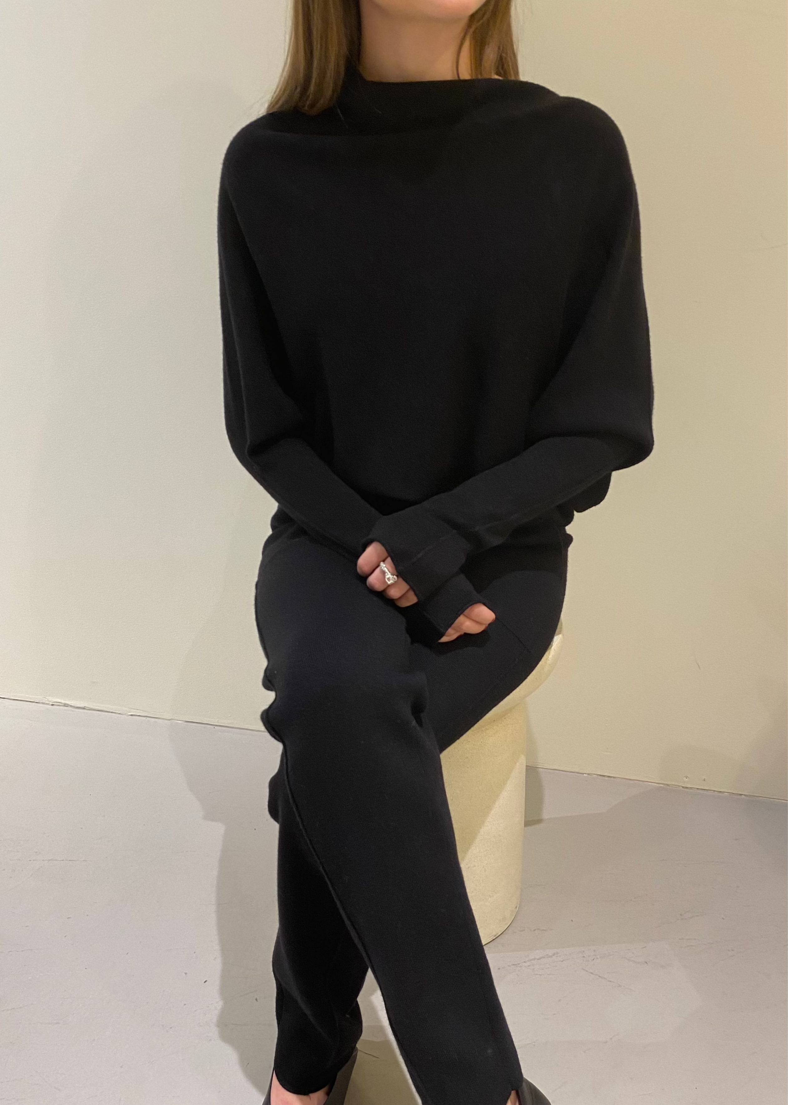 A person wearing a black asymmetric knit top and matching pants, sitting against a neutral background.