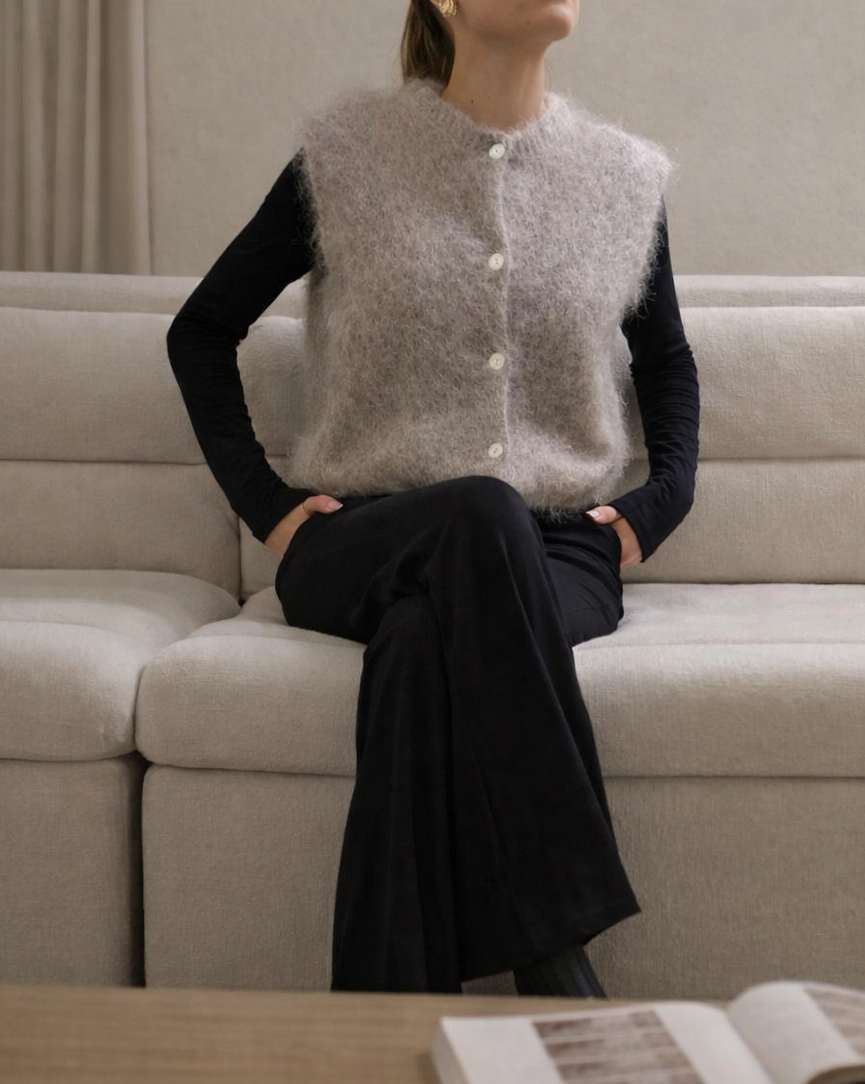 Woman sitting on a beige sofa wearing a gray knitted vest and black pants, with an open book on a wooden table in front of her.