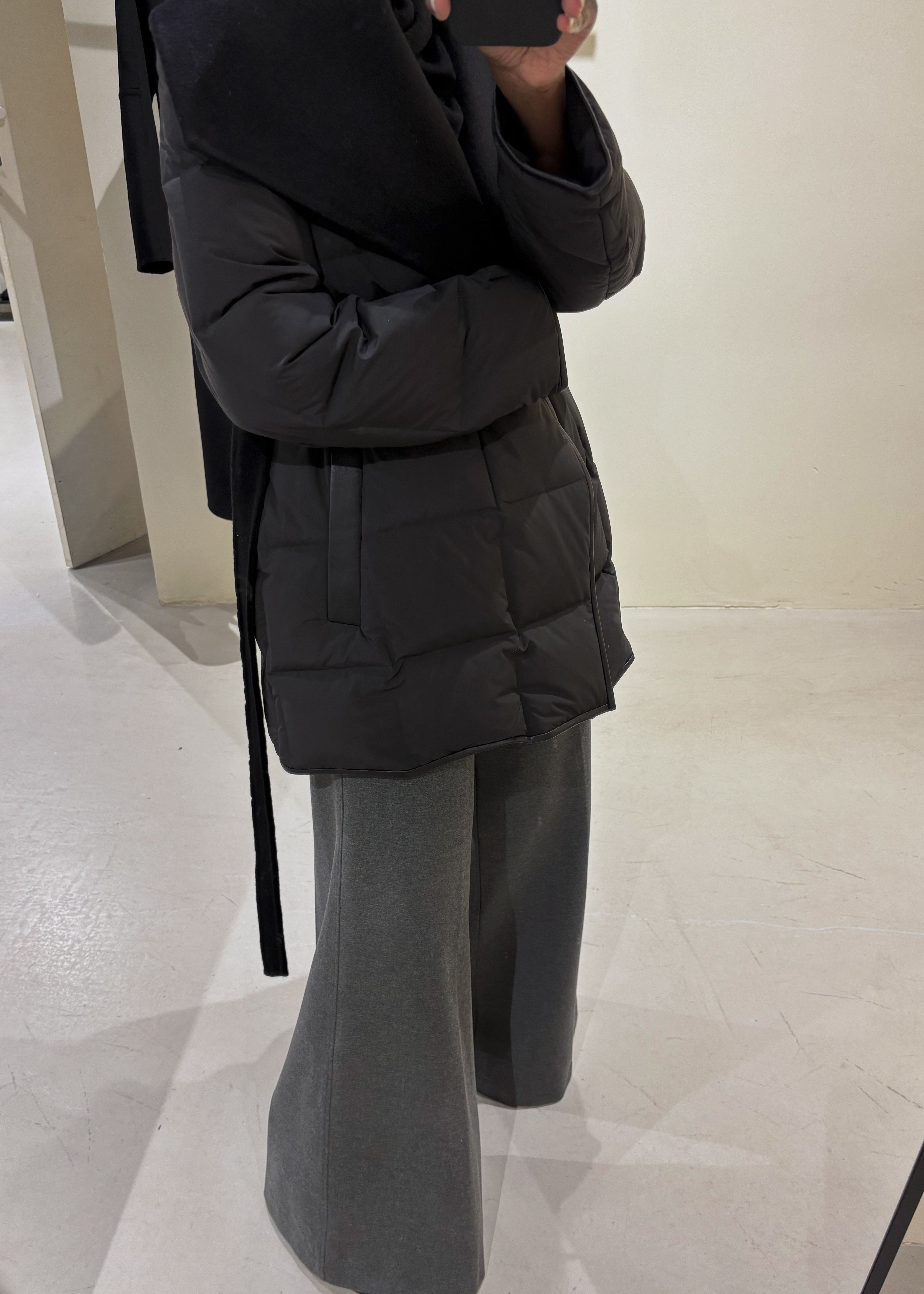 Person wearing a black puffer jacket and gray pants in a minimal indoor setting.