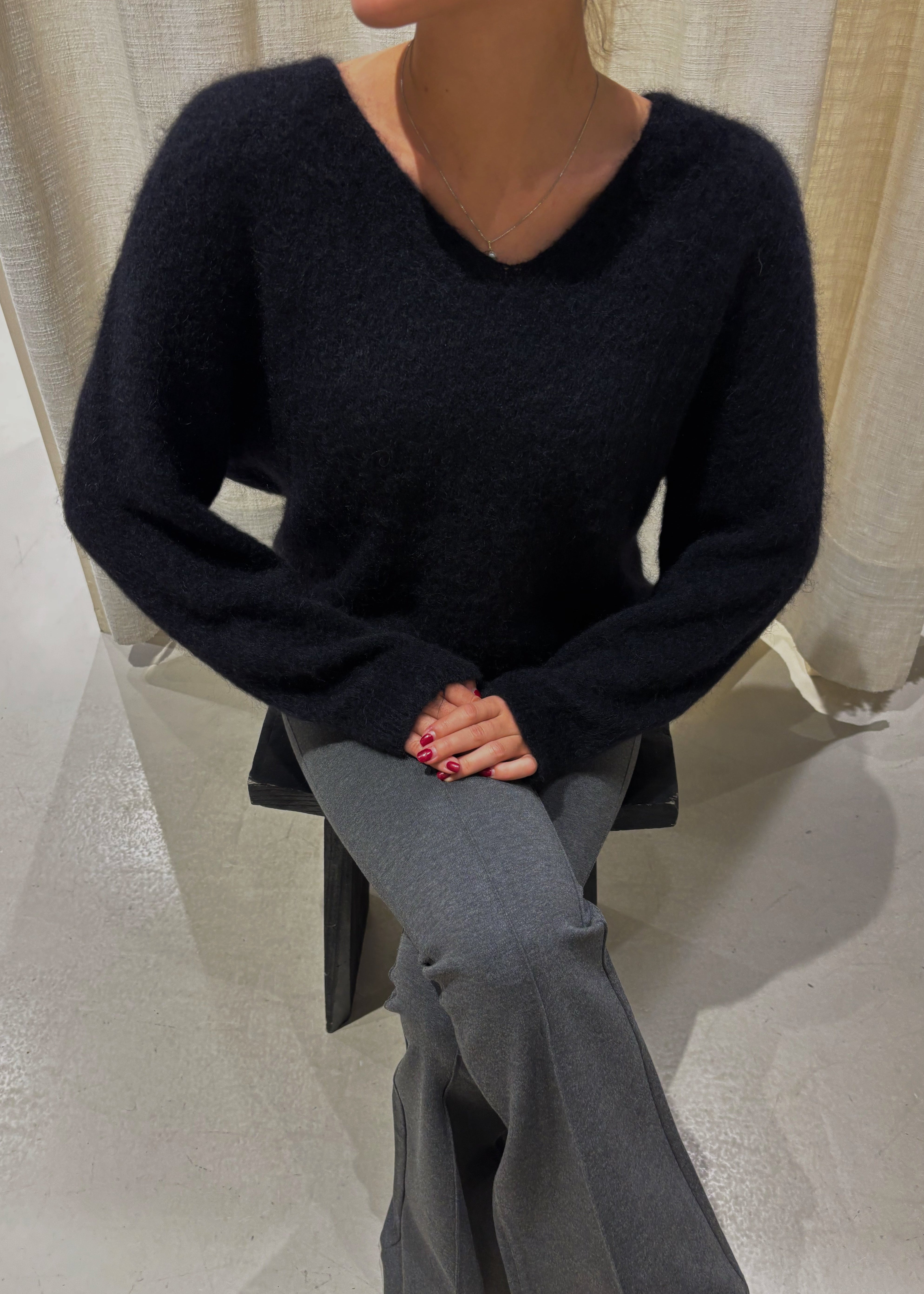 Person wearing a black sweater and gray pants sitting on a white floor with a neutral curtain background